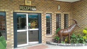 A reception area with a sign indicating reception hours and a dinosaur statue outside. - Queness Inn (Maun)