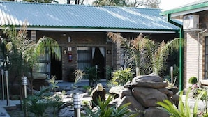 A building with a green roof, a brick facade, and a number '5' sign. There are plants and rocks in the foreground. - Queness Inn (Maun)