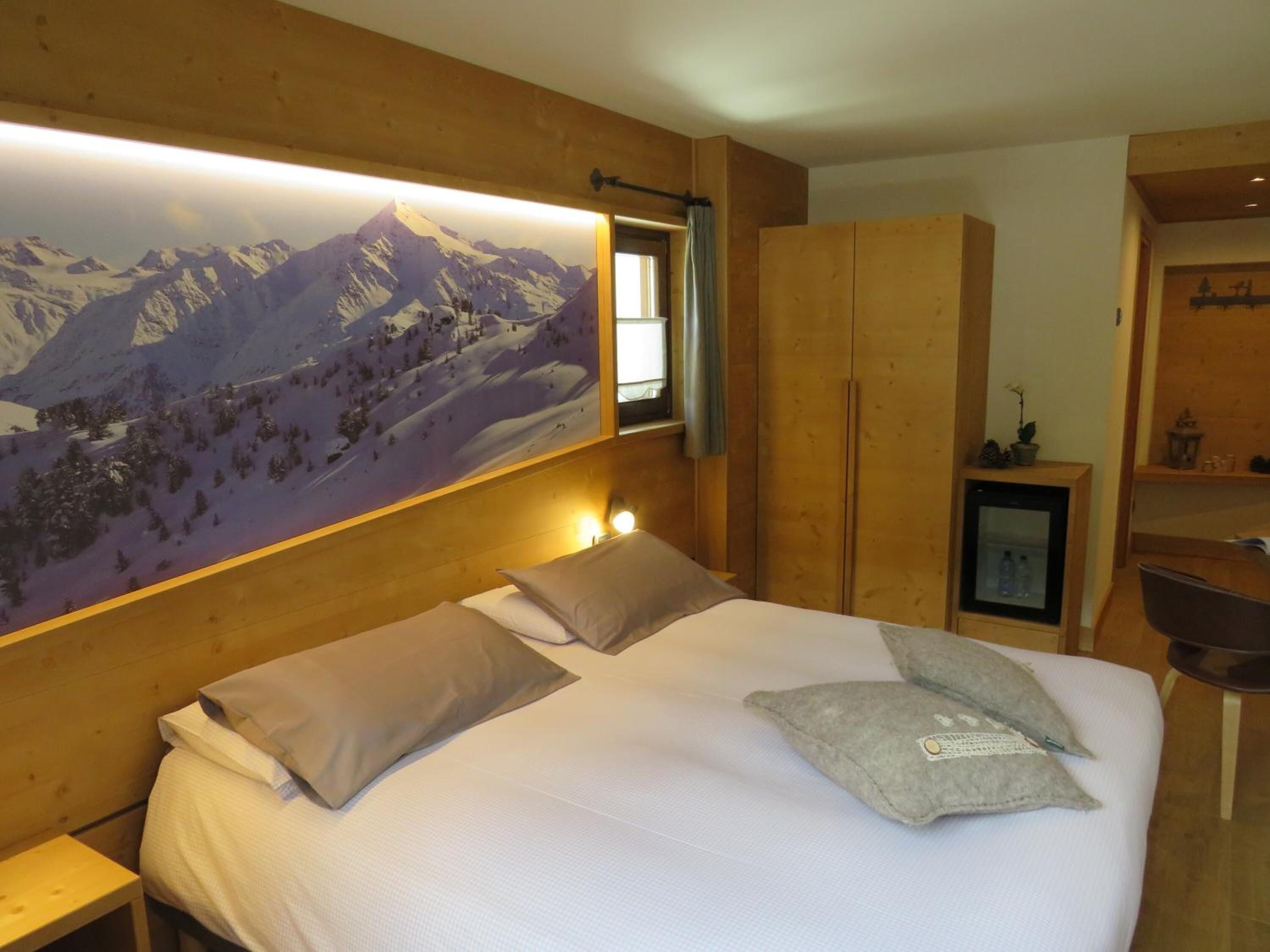 Una camera da letto con un letto grande, una testiera in legno, un quadro appeso con un paesaggio montano, un armadio e un comodino con una lampada.
