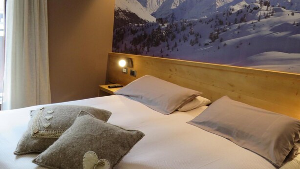 A hotel room with a bed, two pillows, a nightstand, a wall-mounted lamp, and a large mountain view poster. - Hotel Compagnoni (Valfurva)