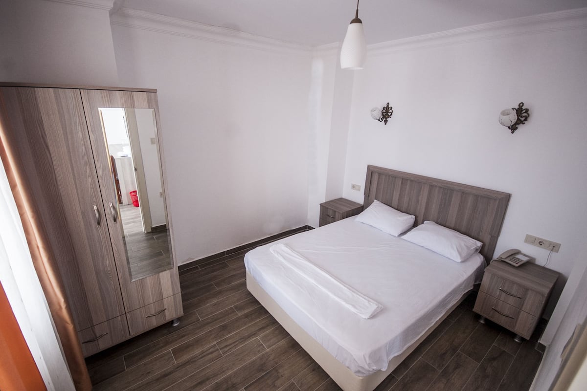 Quarto com guarda-roupa de madeira, cama com roupa de cama branca, mesa de cabeceira com telefone e criado-mudo.