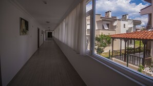 Un corredor largo y estrecho con suelo de madera y paredes blancas, que conduce a un balcón con vistas a edificios vecinos.