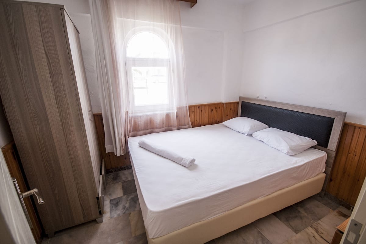 Quarto com guarda-roupa de madeira, cama com roupa de cama branca e janela com cortinas leves.