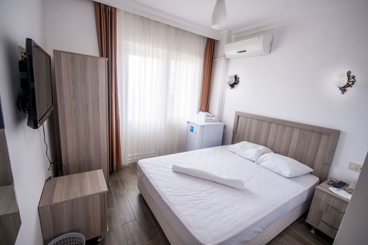 Quarto de hotel com cama, televisão, pequena escrivaninha, guarda-roupa e ar condicionado.