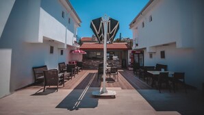 Un área de café al aire libre con mesas y sillas, un refrigerador de Coca-Cola y una pared de piedra al fondo.