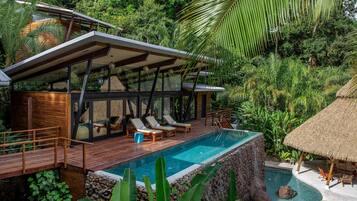 Una villa moderna con piscina, terraza de madera y zona de estar con techo de paja, rodeada de exuberante vegetación.