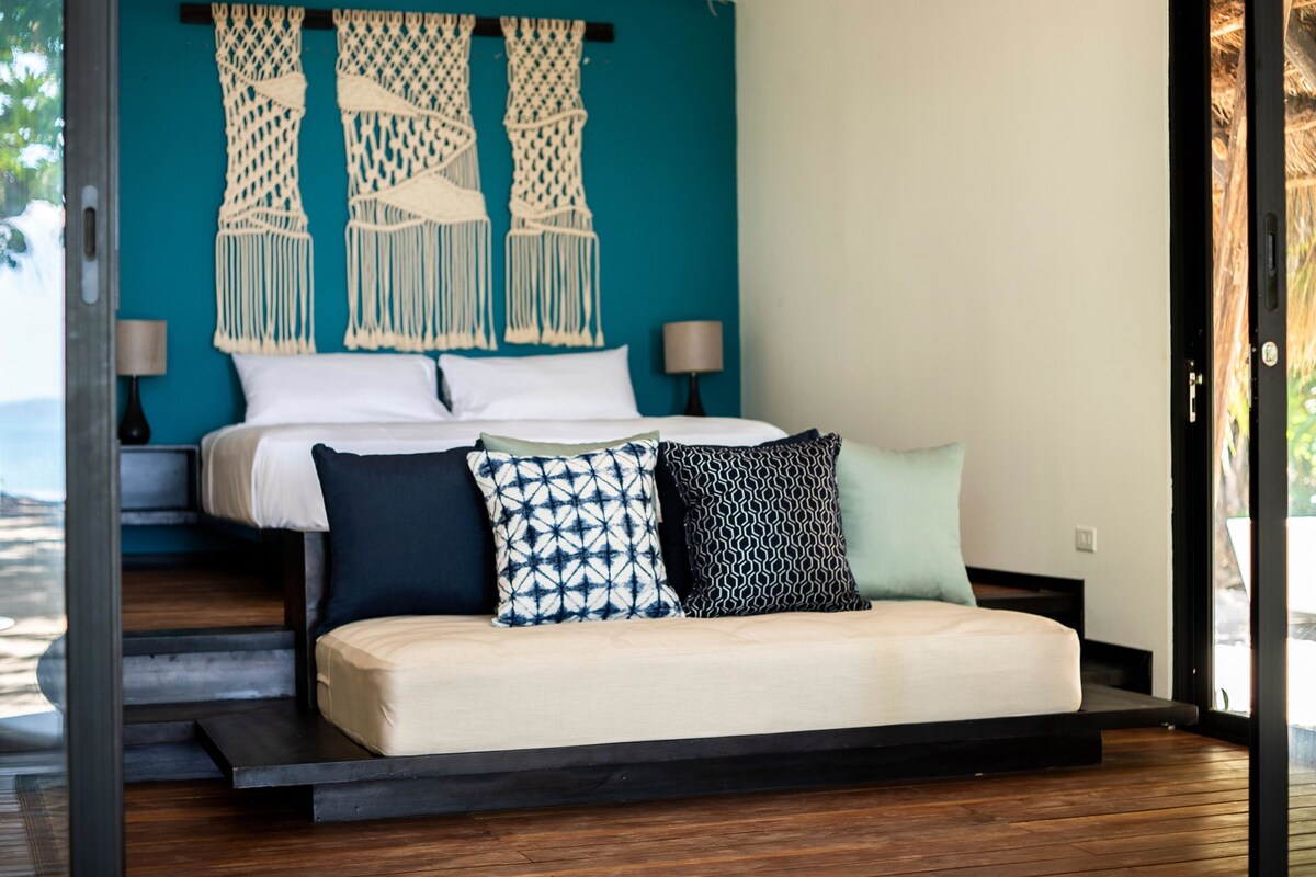 A bed with a white mattress, multiple pillows, and macrame wall hangings.