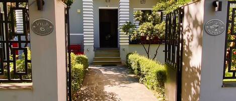 Una entrada de hotel con un camino empedrado que lleva al edificio principal, un letrero que indica el nombre del hotel y un balcón con barandilla.