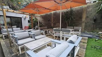 Un patio exterior con mobiliario gris y blanco, que incluye sofás, sillas y mesas, bajo un amplio paraguas naranja.
