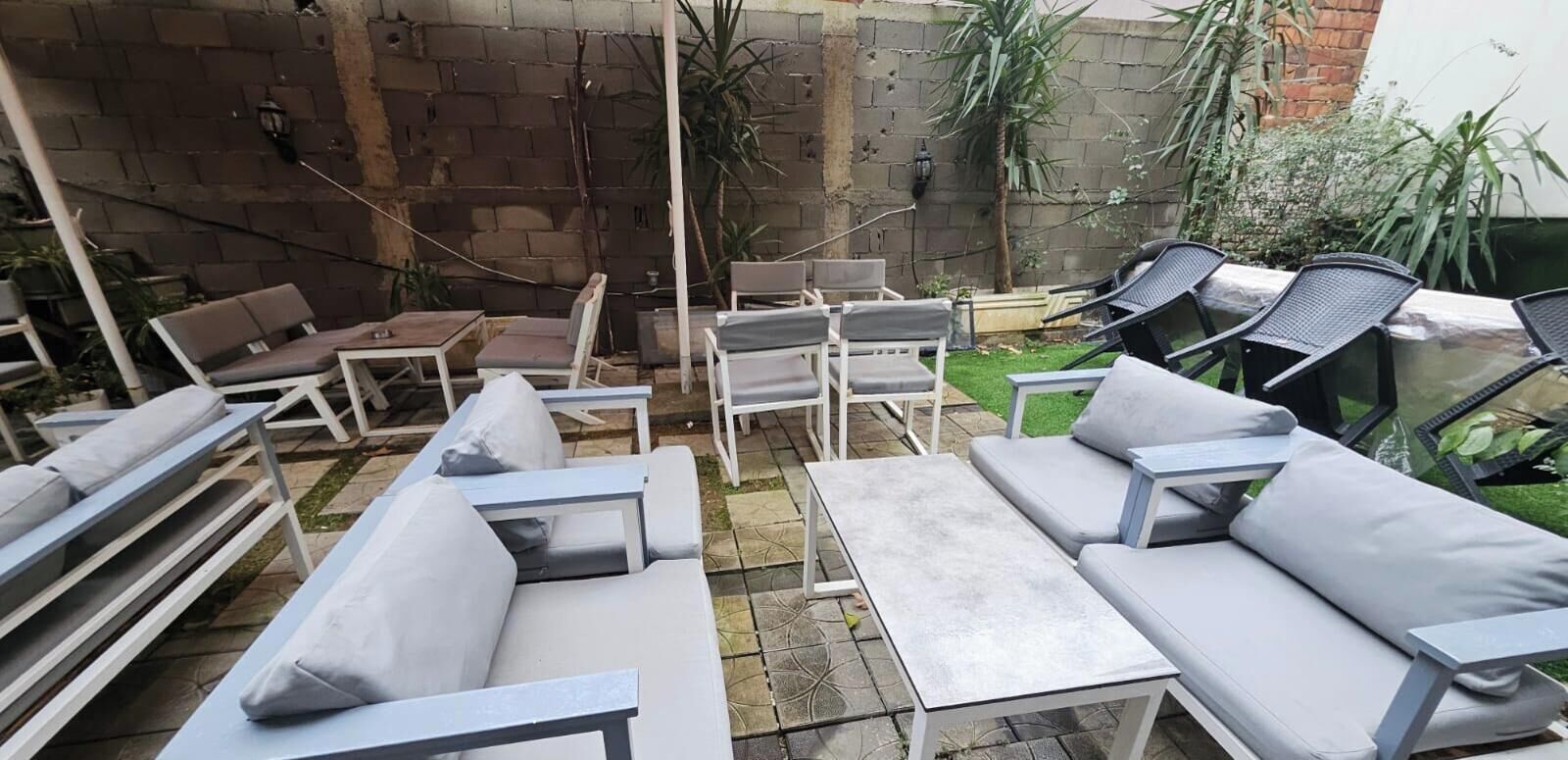Un patio amueblado al aire libre con sofás, sillas y una mesa, frente a una pared de ladrillo con plantas y un pequeño jardín.