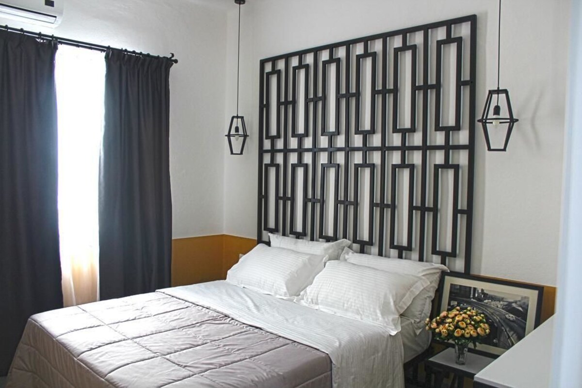 Un dormitorio con cama, un panel geométrico en la pared, una mesita de noche con un jarrón y un marco de cuadro.