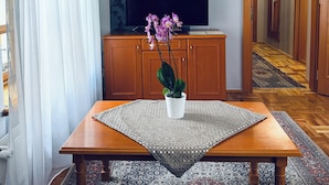 Una sala de estar con una mesa de madera, una orquídea en maceta, un televisor de pantalla plana sobre un mueble, papel tapiz estampado y un pasillo con piso de madera.