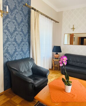Una sala de estar con un sofá de cuero negro, una mesa de centro de madera, una orquídea en maceta, una lámpara y un espejo en la pared.