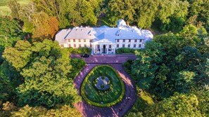 Une vue aérienne d’un grand bâtiment blanc à la façade symétrique, entouré d’une végétation luxuriante et d’un jardin circulaire avec une fontaine centrale.