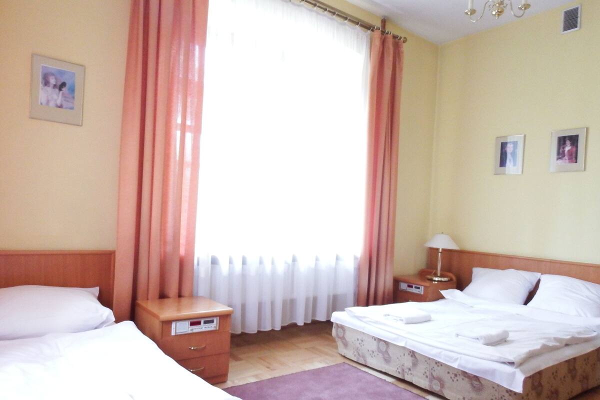 Habitación de hotel con dos camas, una mesita de noche con lámpara, un pequeño aparador y una ventana con cortinas.