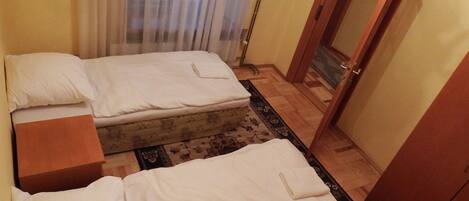 Una habitación con dos camas, piso de madera, una puerta y un ventanal con cortinas.
