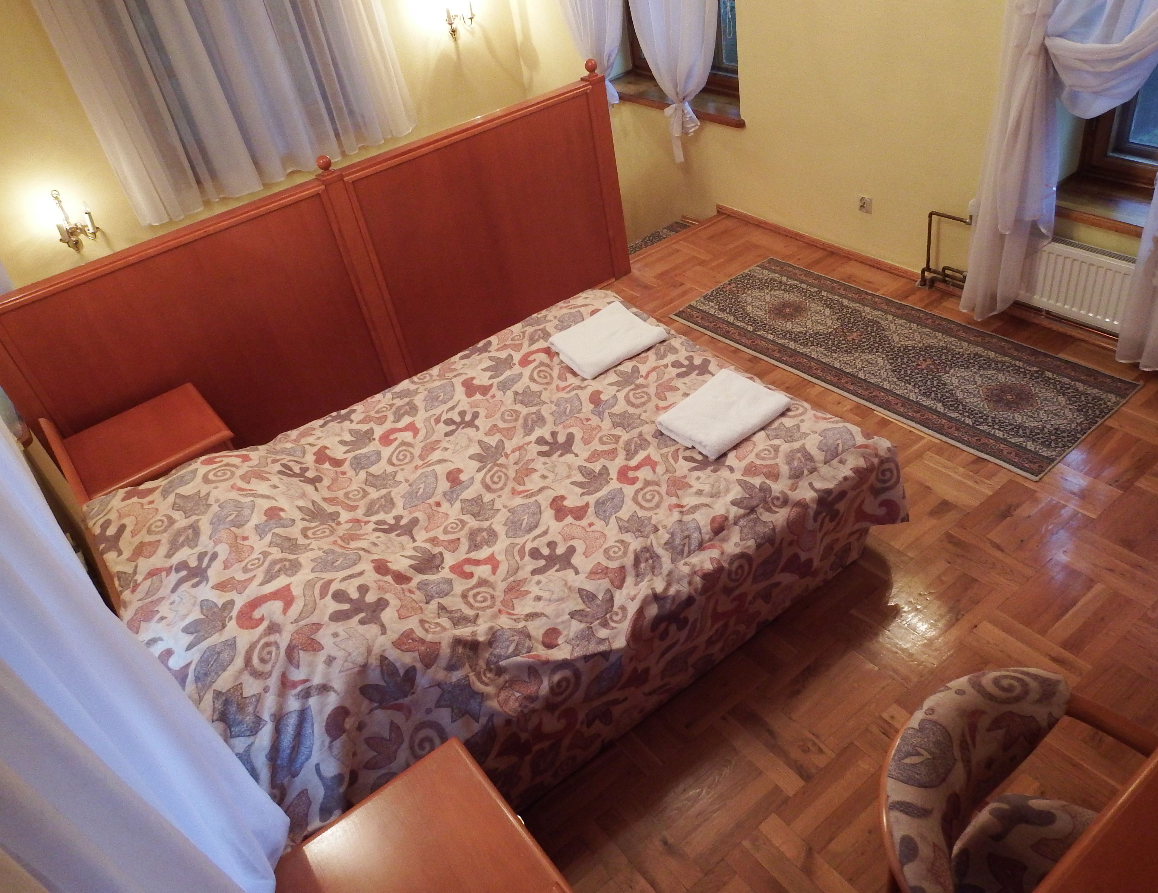 Camera da letto con testiera in legno, copripiumino decorato, due asciugamani sul letto, pavimento in legno, tappeto e finestra con tende.