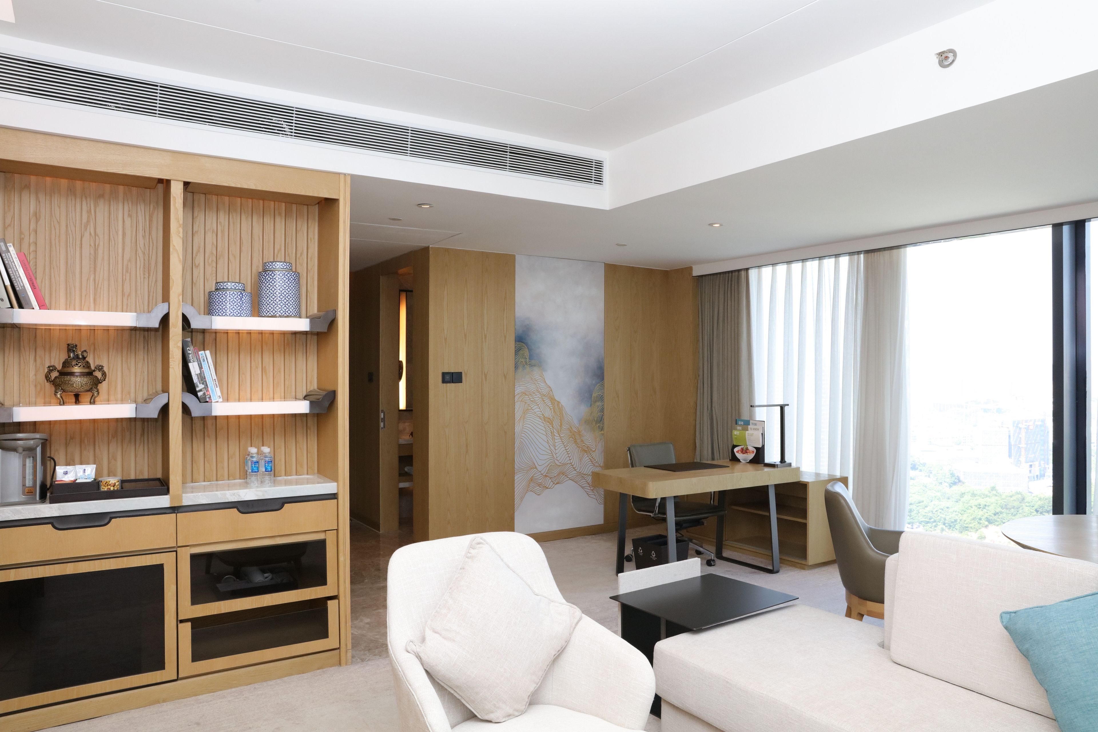 Een moderne hotelkamer met een houten kast, een bank, een eettafel en een groot raam met gordijnen.