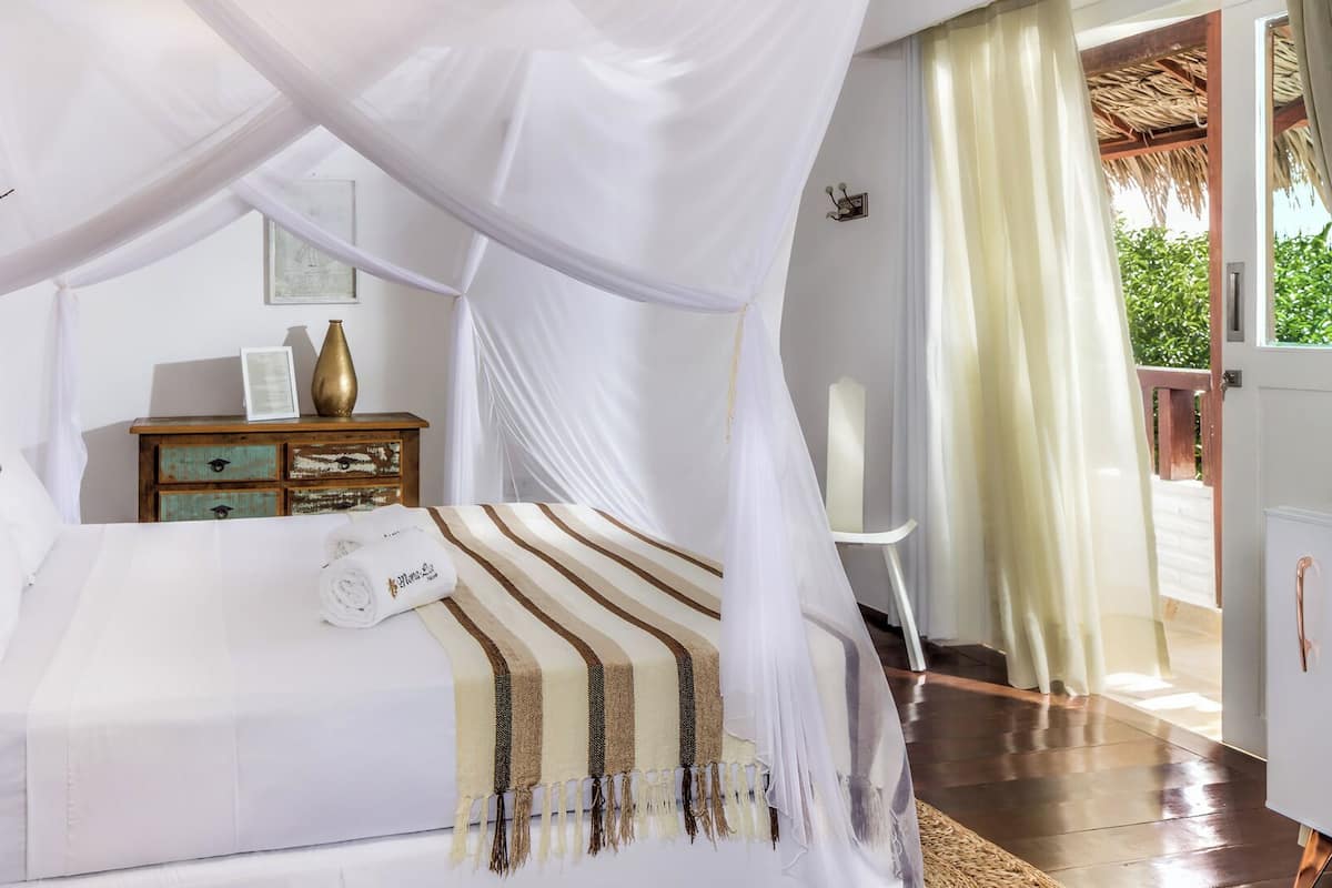 A bedroom with a canopy bed, a wooden nightstand with a vase, and a view of greenery outside.