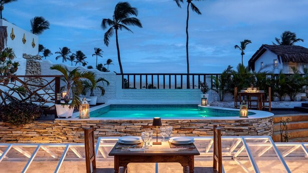 A pool with a stone wall, palm trees, and a dining area with a table set for two. - Pousada Monalisa (Jijoca de Jericoacoara)