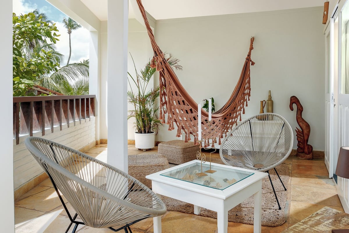 A sunlit patio with two wicker chairs, a glass-top table, a hammock, and potted plants.