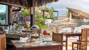 A dining area with a wooden table set for breakfast, surrounded by chairs, with a view of a stone wall and tropical plants. - Pousada Monalisa (Jijoca de Jericoacoara)