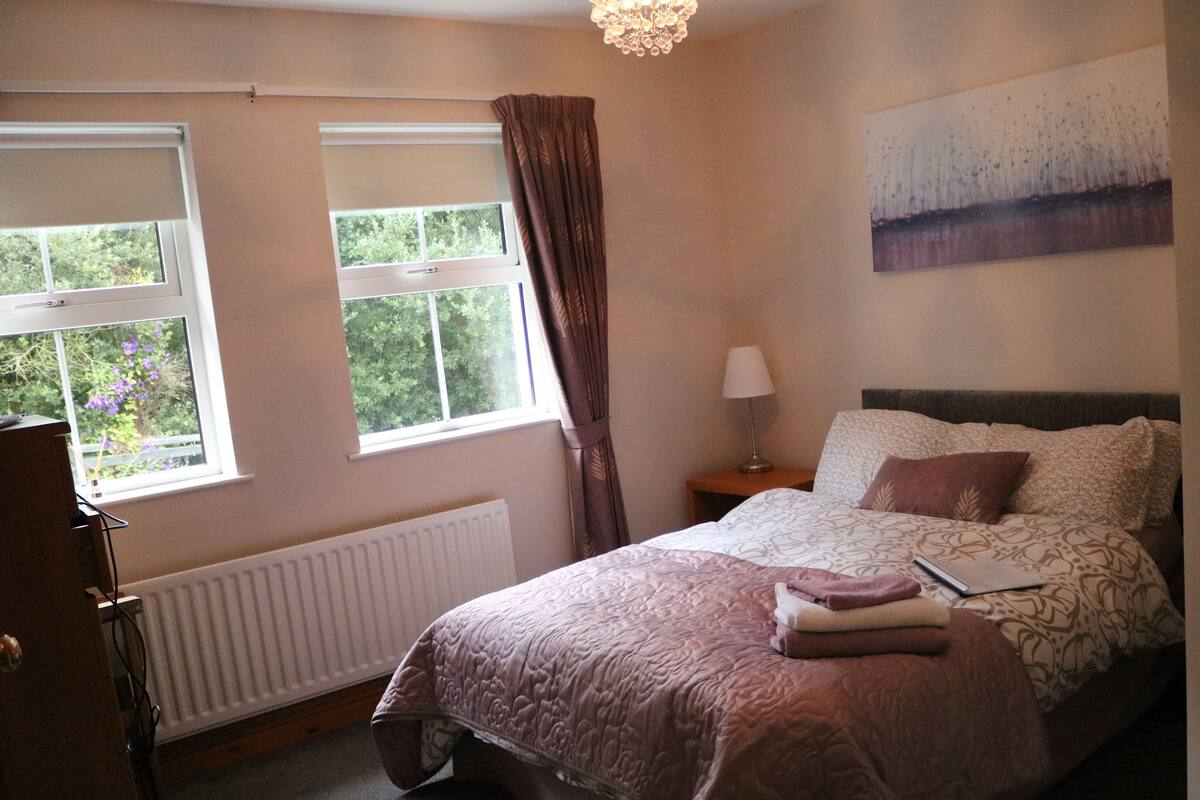 A bedroom with a bed, a radiator, a window with curtains, a lamp, and a painting on the wall.