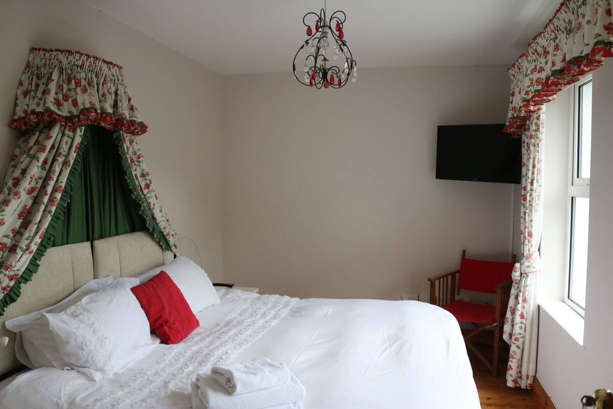 A neatly made bed with white linens and a red pillow, a wooden chair, a television mounted on the wall, and a window with floral curtains.