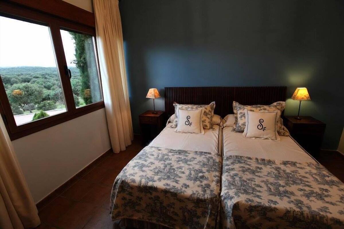 A double bed with floral bedding, two bedside lamps, and a view of greenery through a window.