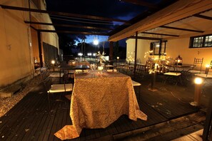 An outdoor dining area with tables and chairs, covered by a pergola, and illuminated by string lights. - La Senda de los Enebros (Higuera de las Duenas)