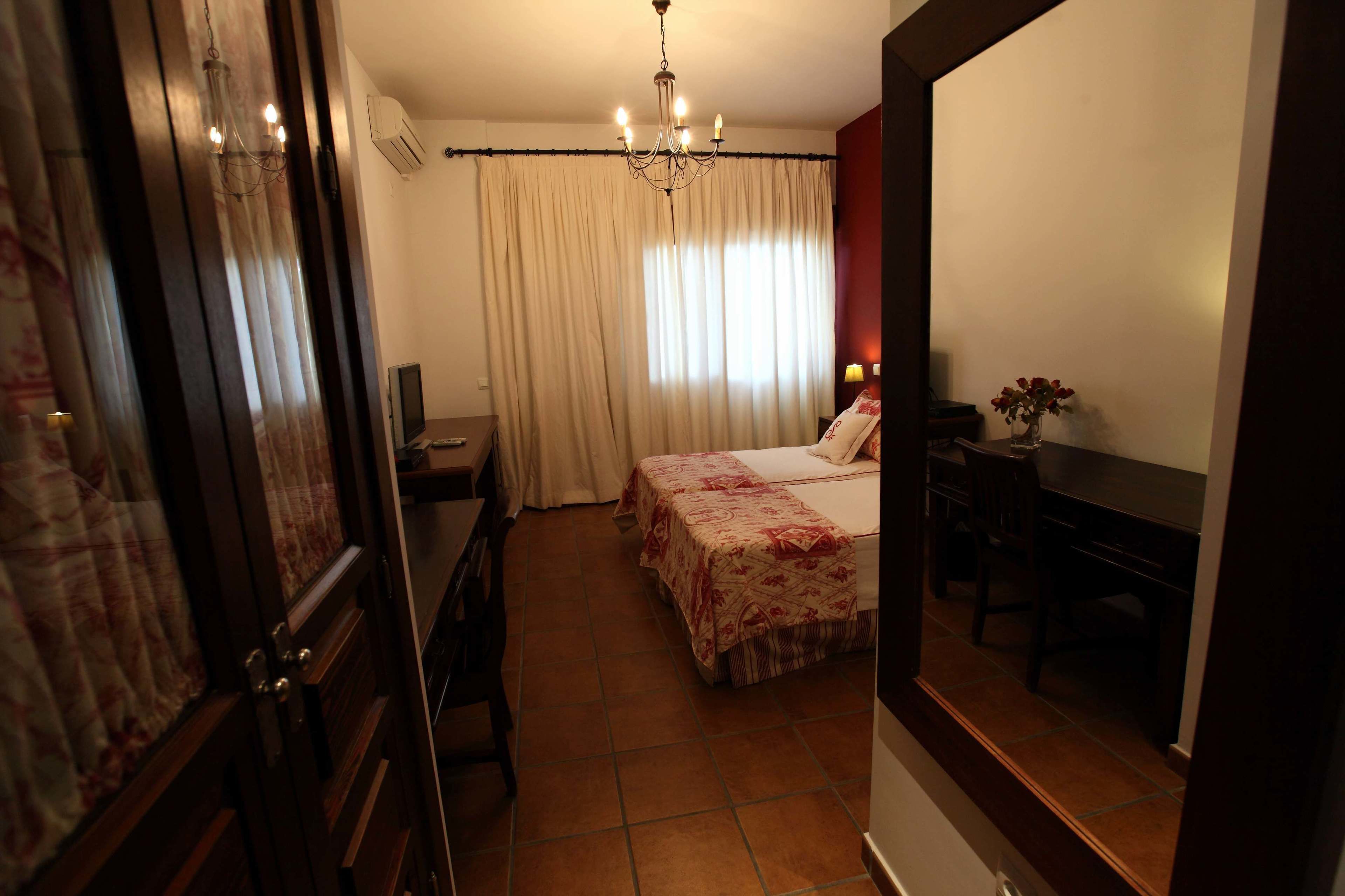 A bedroom with a bed, a desk, a chair, a TV, and a chandelier.
