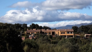 A row of modern, two-story buildings with a uniform architectural style, surrounded by lush greenery and mountains in the background. - La Senda de los Enebros (Higuera de las Duenas)