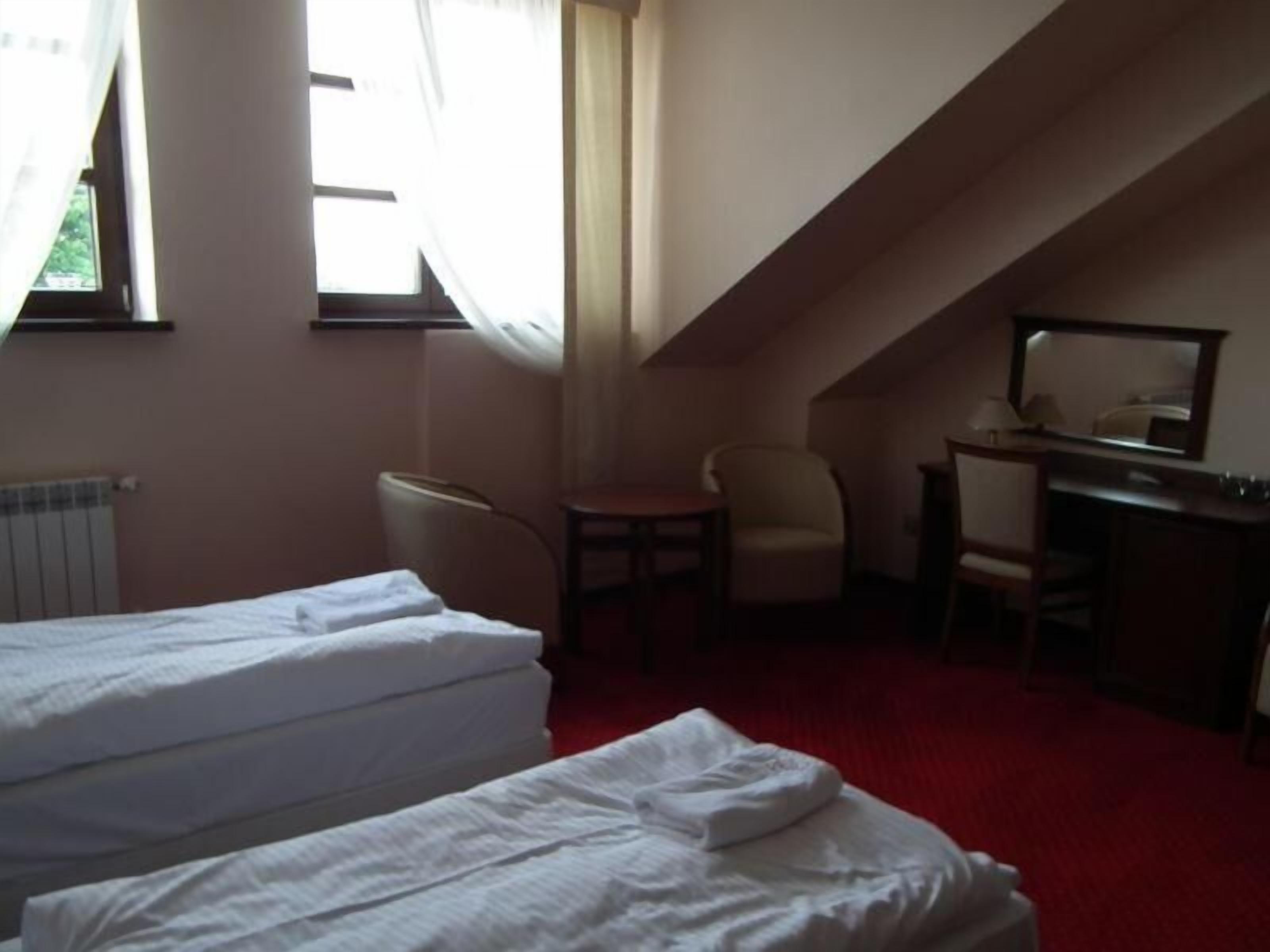A small, dimly lit room with two beds, a desk, a chair, and a window with curtains.