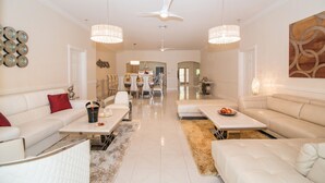 A spacious living room with a high ceiling, white sofas, a dining area, and a chandelier.