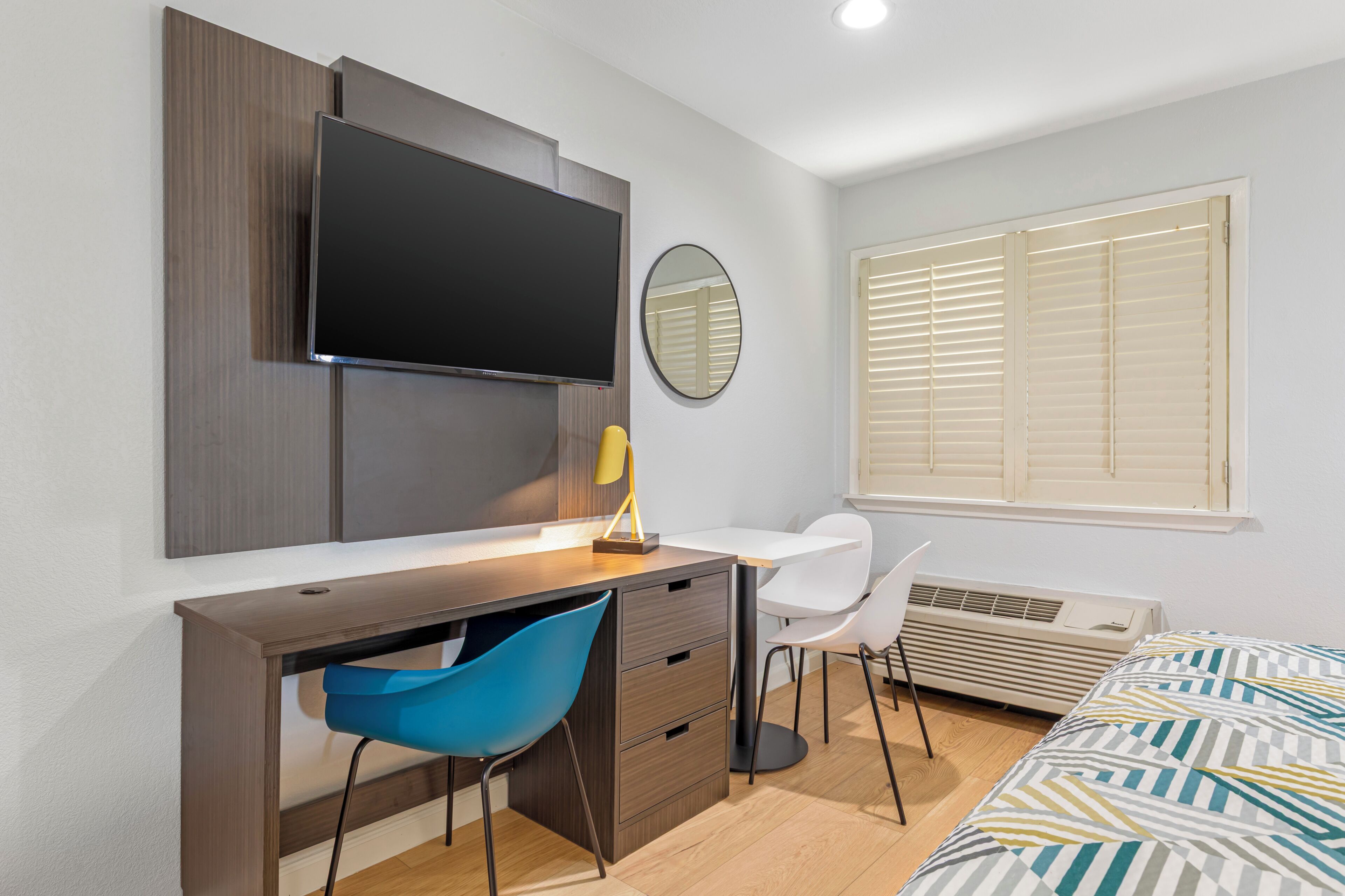 A modern bedroom with a bed, a desk, a chair, a TV, and a window with blinds.