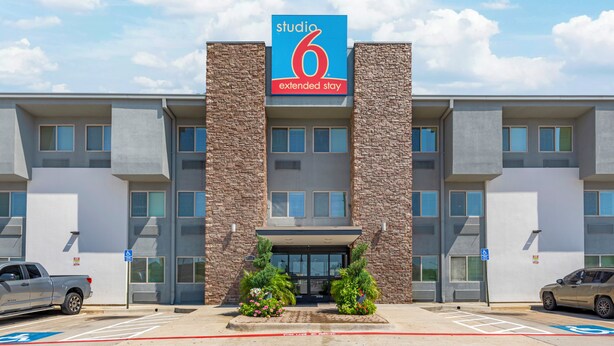 Un hotel moderno con un llamativo letrero con el número '6', un estacionamiento y una zona de acceso para vehículos de emergencia.
