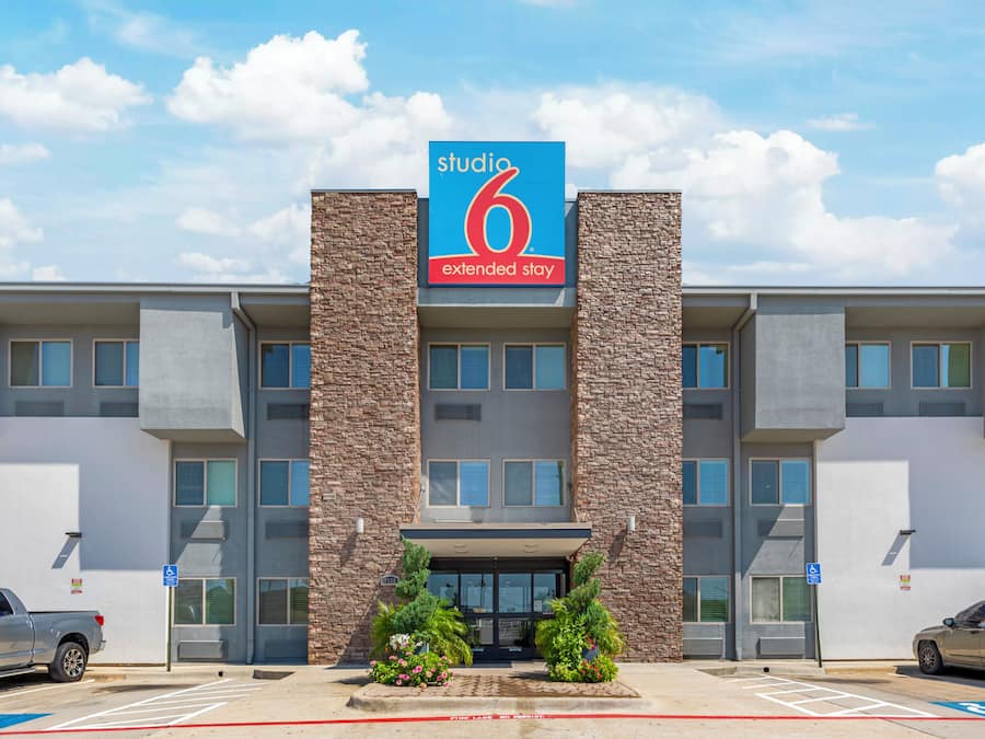 Un hotel moderno con un llamativo letrero con el número '6', un estacionamiento y una zona de acceso para vehículos de emergencia.