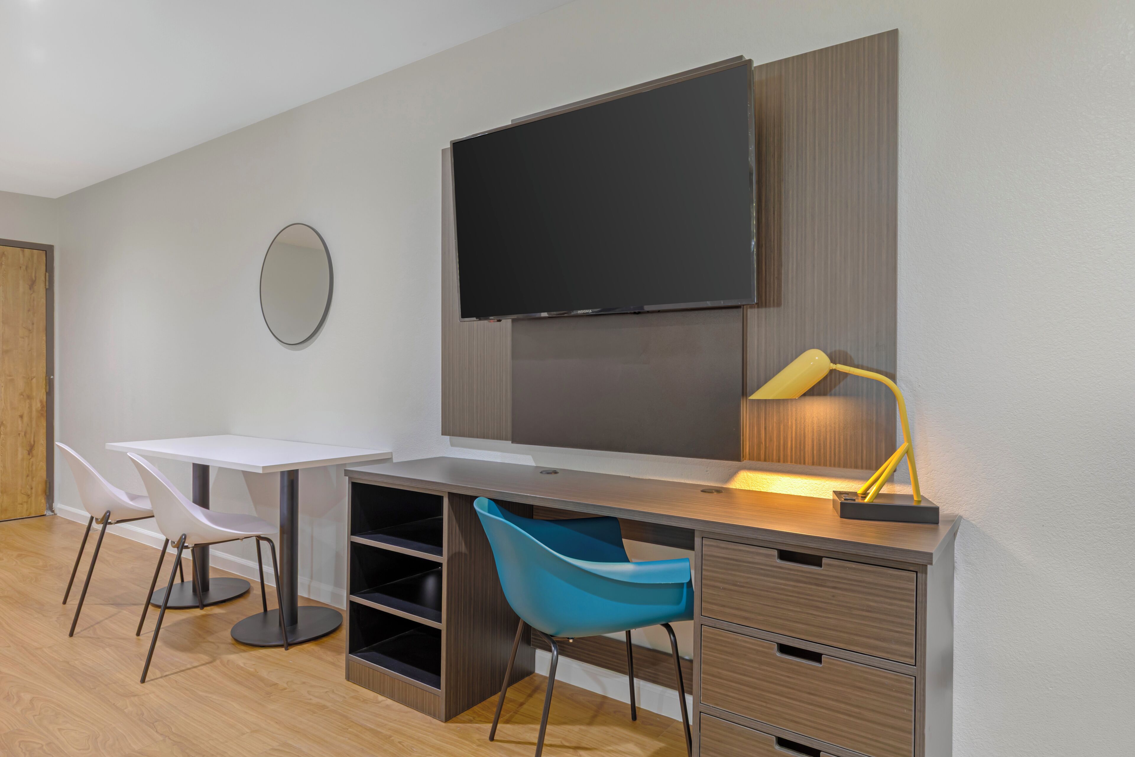 Una habitación moderna con un televisor de pantalla plana montado en la pared, un escritorio con una silla, una silla azul y una lámpara de escritorio.