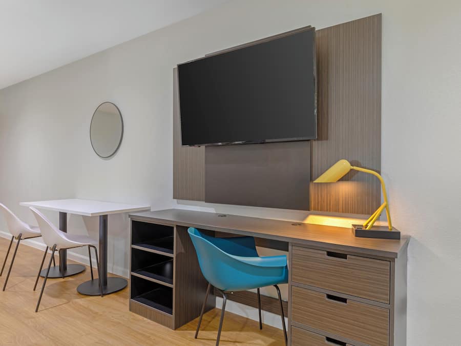 A modern room with a flat-screen TV mounted on the wall, a desk with a chair, a blue chair, and a desk lamp.