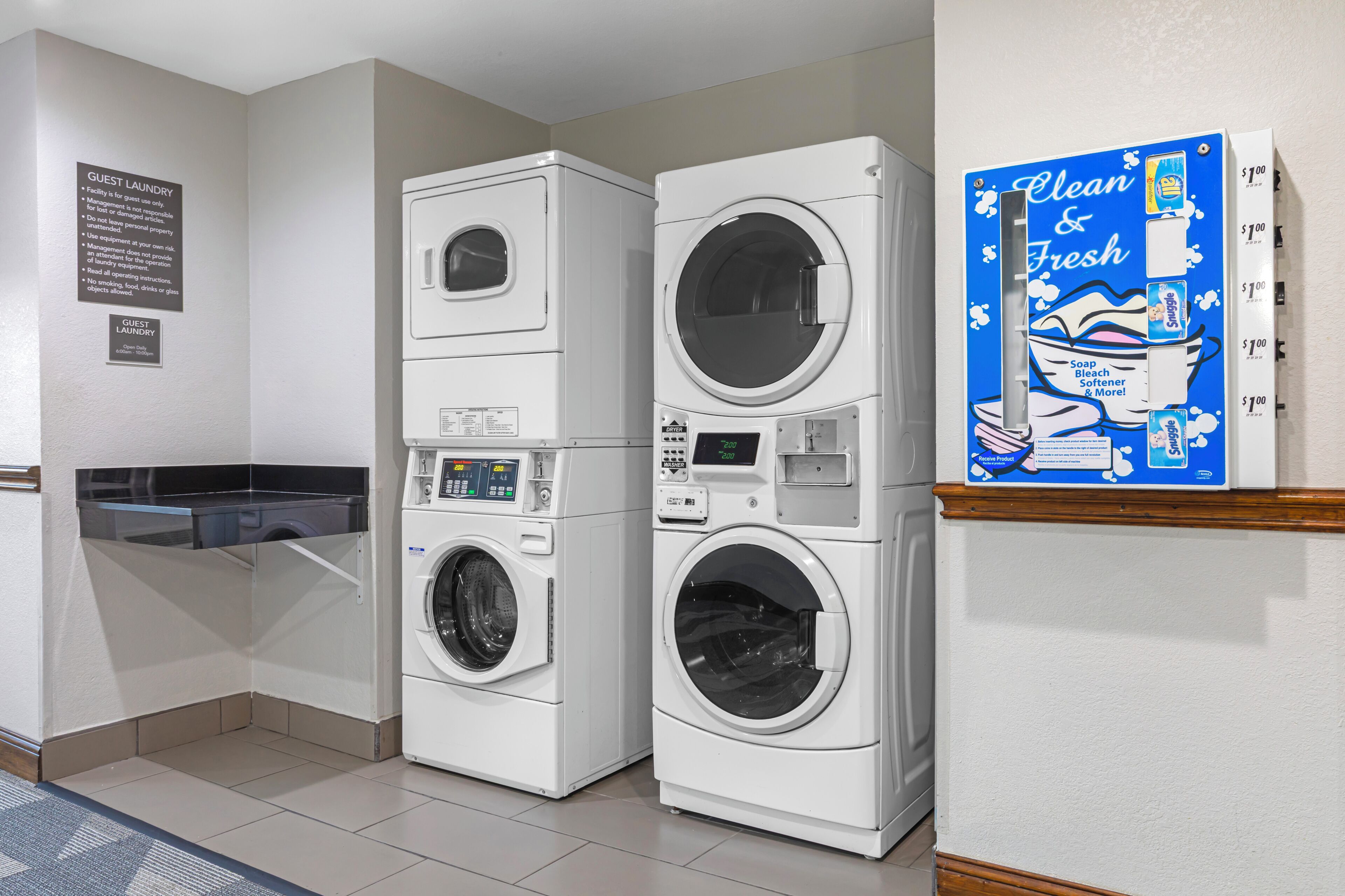 A laundry facility with stacked washers and dryers, a sign indicating guest laundry use, and a promotional sign for a cleaning product.