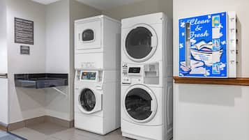 A laundry facility with stacked washers and dryers, a sign indicating guest laundry use, and a promotional sign for a cleaning product.