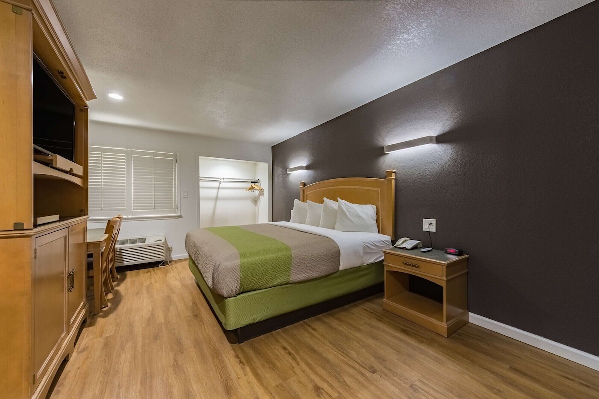 A hotel room with a wooden bed, a bedside table with a phone and charger, a wardrobe, and a window with blinds.