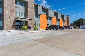 A multi-story building with a parking lot and several cars. - Studio 6 Plano, TX (Plano)