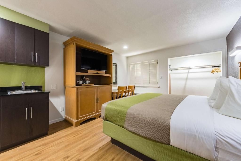 A compact hotel room with a bed, a television, a kitchenette, and a window with blinds.