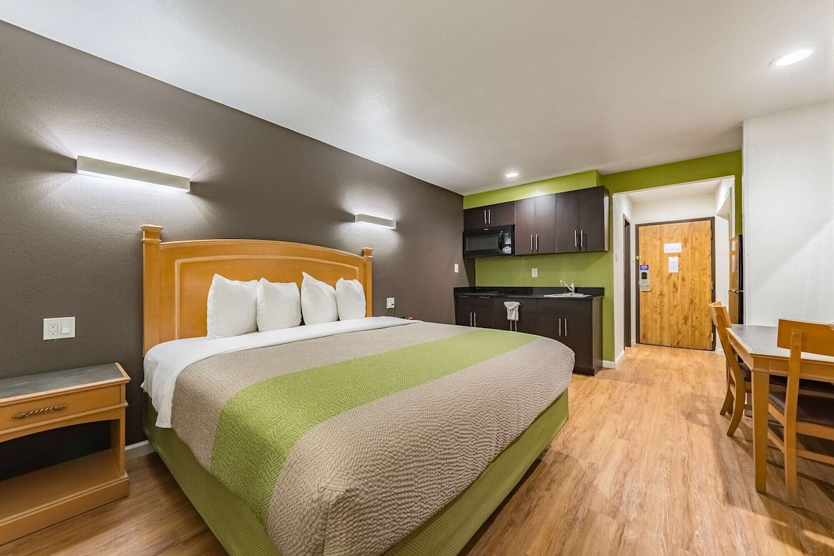 A compact hotel room with a bed, bedside table, a small kitchenette, and a dining area.