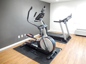 Dos bicicletas estáticas sobre tapetes negros en una habitación con suelo de madera y una pared gris.