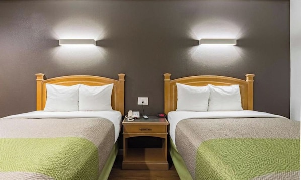A hotel room with two beds, each with a wooden headboard and white bedding. There is a nightstand between the beds with a phone and a small lamp. - Studio 6 Plano, TX (Plano)