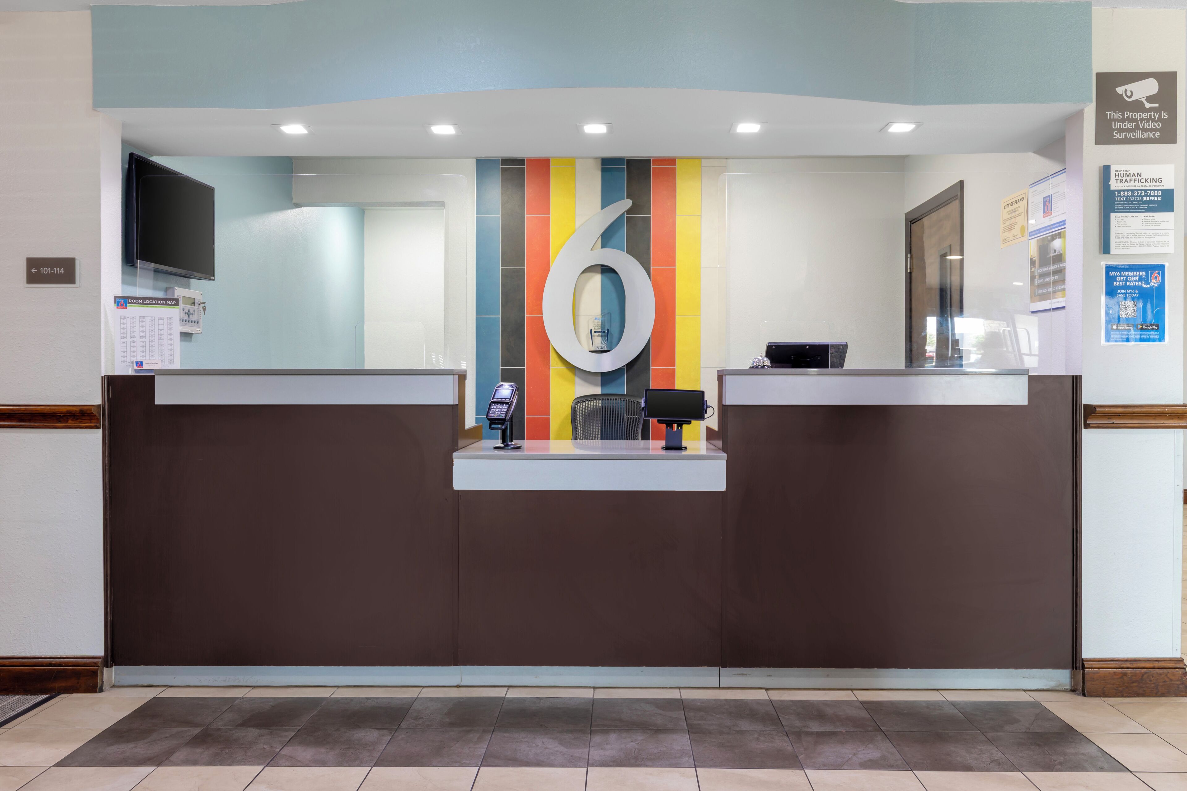 Un mostrador de recepción con un gran número '6' en la pared, una pantalla digital y una cámara de seguridad.