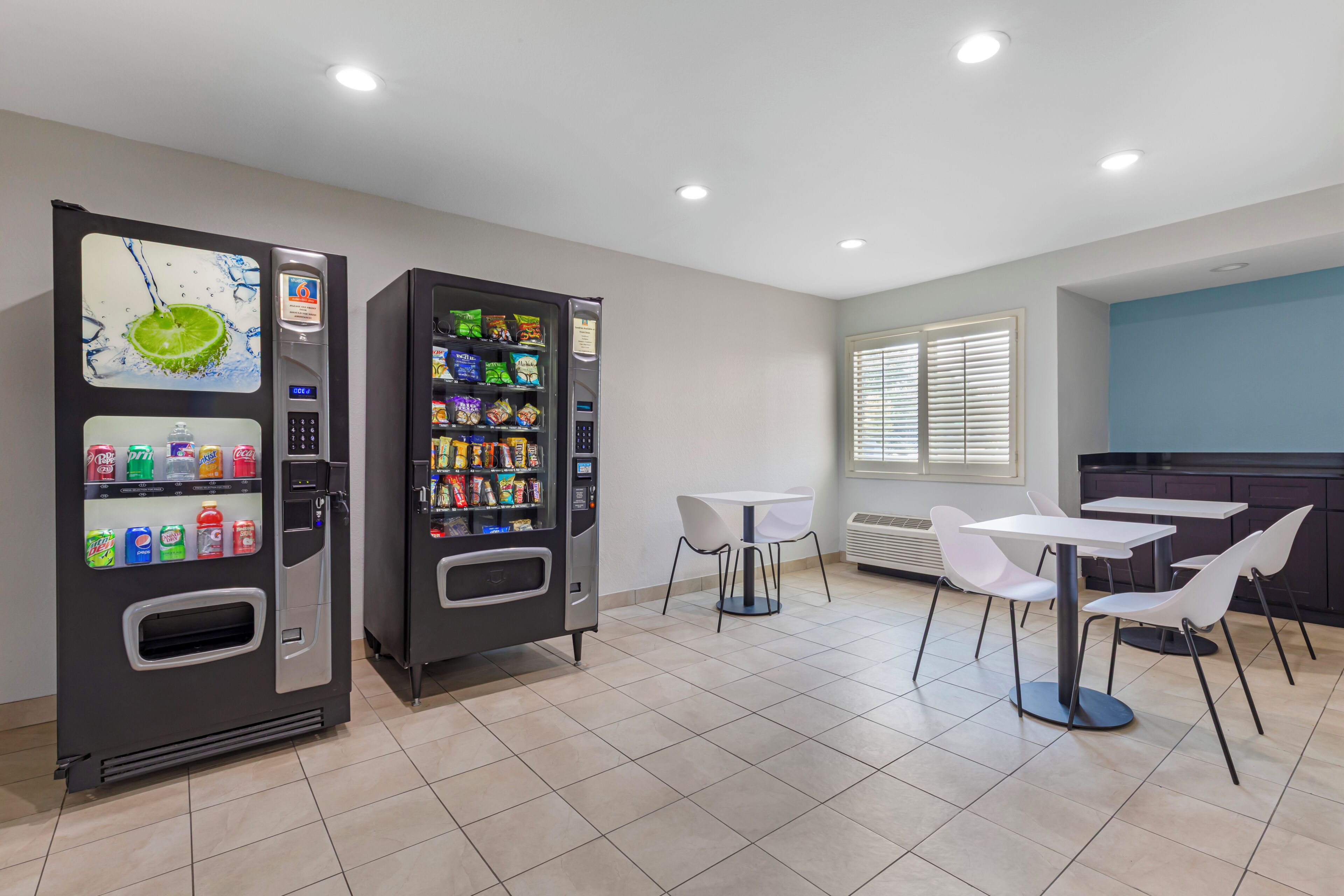 A clean, modern room with two vending machines, a table with chairs, and a window with blinds.