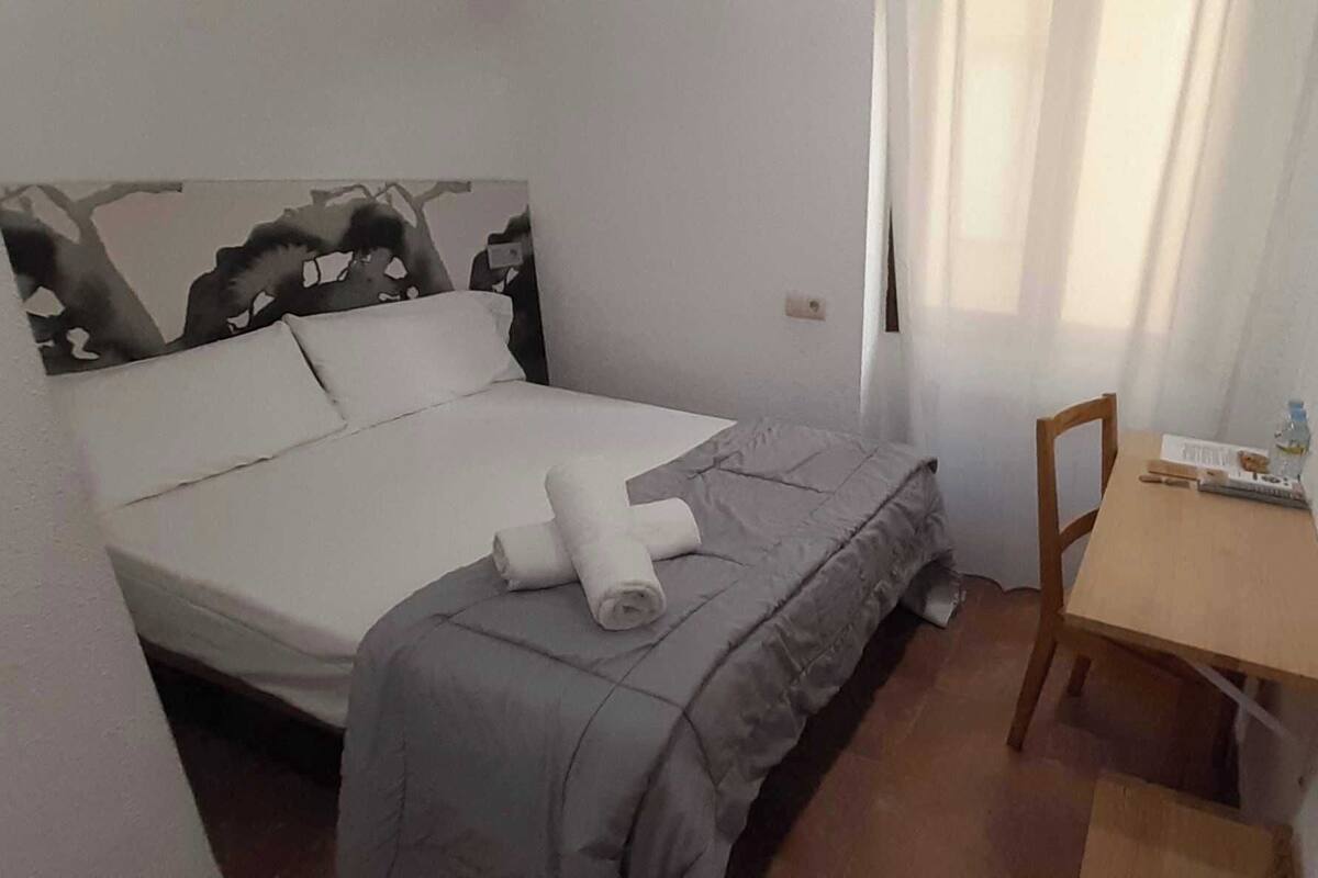 A small, neatly arranged bedroom with a bed, a chair, a table, and a window with curtains.